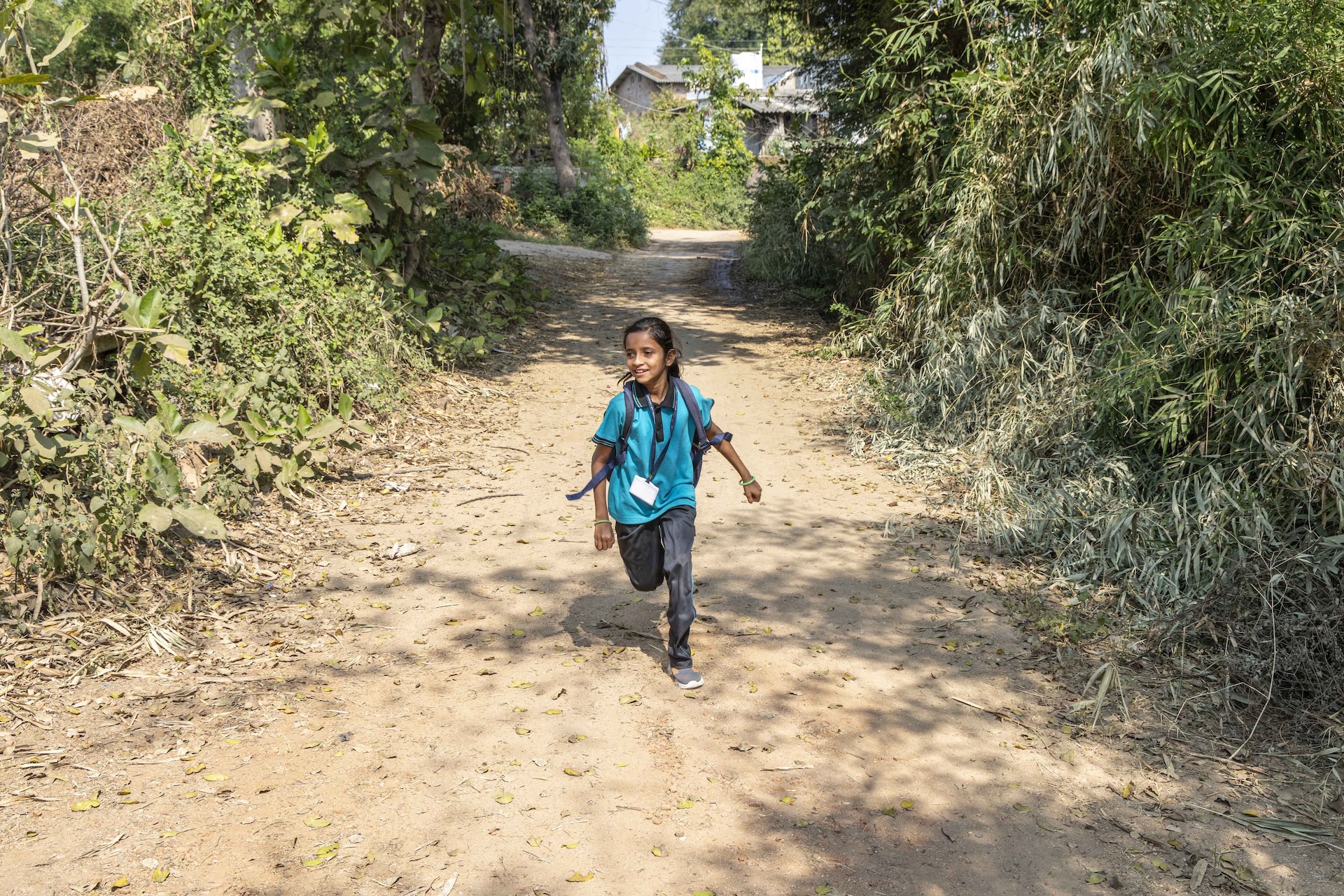 Suhaniben Kishanbhai (10) runs towards the camera on a beige muddy lane. Her straight black hair is combed back and tied. She wears ash-grey trousers and a dark sky blue half-sleeved knitted shirt. The shirt has an ash-grey collar and the edges of the sleeves are also ash-grey. A jade-green bangle encircles each wrist. The broad ash-grey straps of her backpack run down from her neck and shoulders to her armpits. Bordering both sides of the lane are trees, saplings, and bushes.