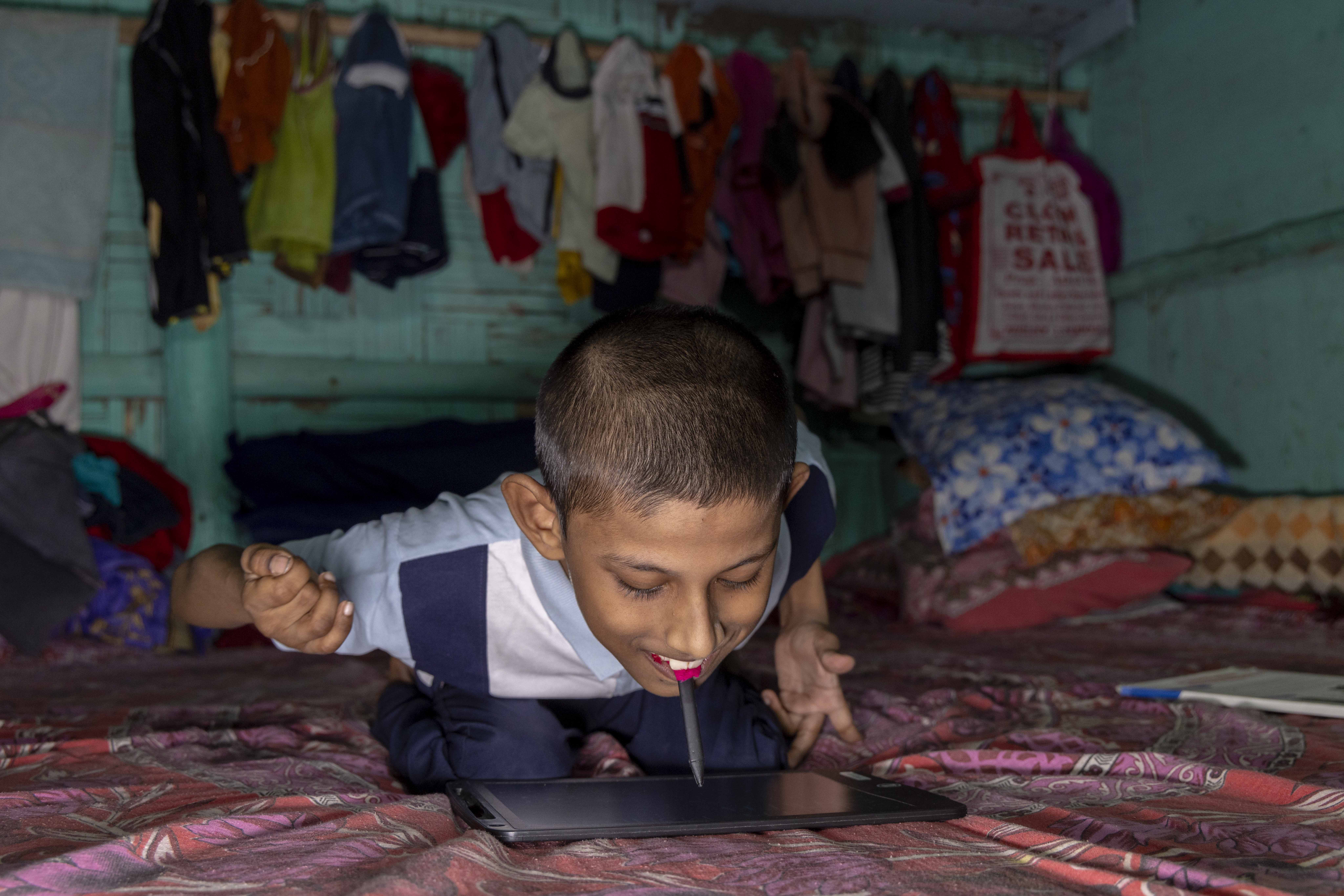 Sumith Mondal (12) sits on a bed, crouching over a black writing tablet with an LCD screen. He holds the black stylus in his mouth. His hair is cropped close with the scalp showing through. He wears a pale blue knitted shirt and the bedspread is printed in a rose-pink and white design.