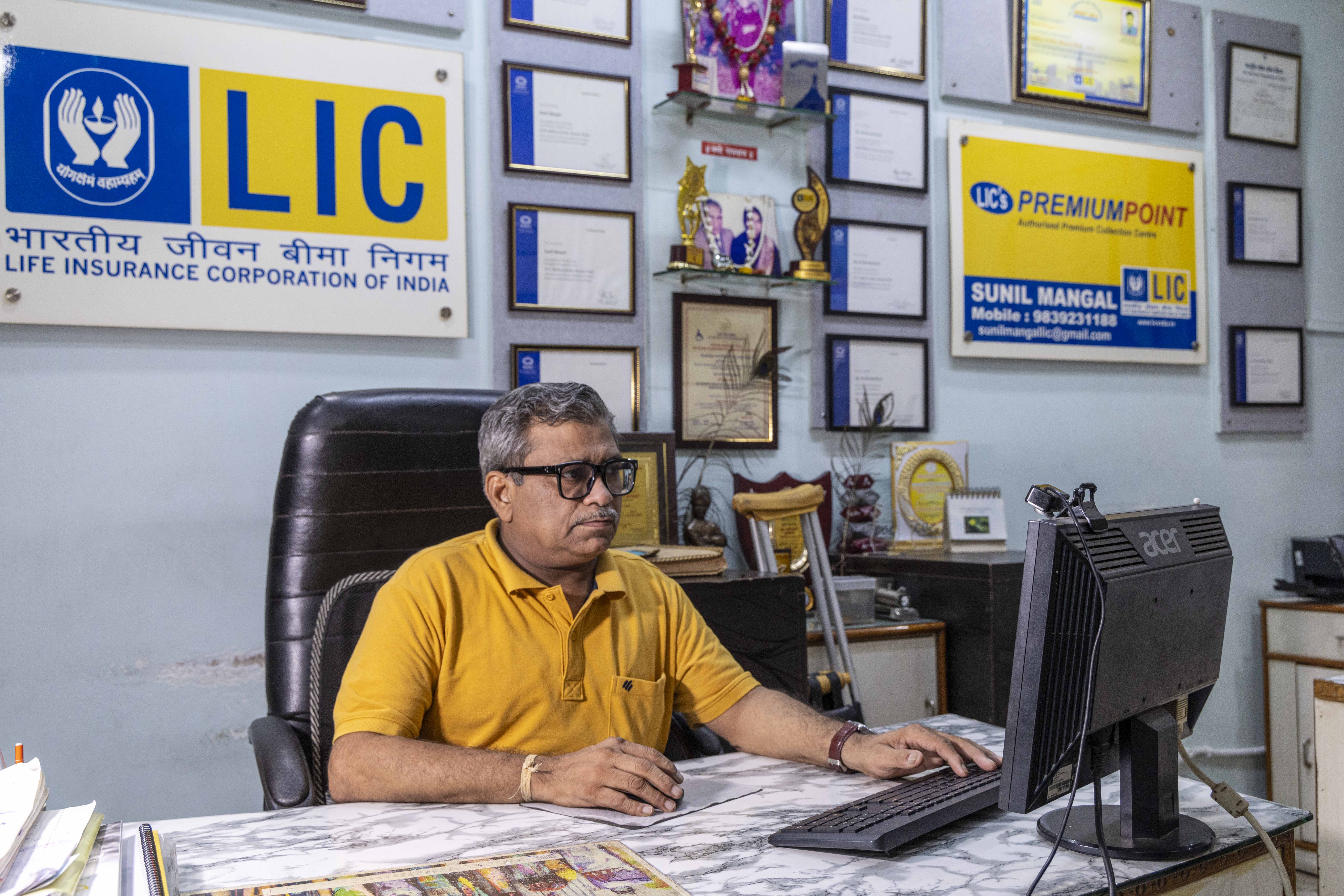 Sunil Mangal (53), wearing a mustard-yellow half-sleeved knitted shirt, sits behind a table, looking at the screen of a black desktop computer with his left hand on the black keyboard. A narrow cream band consisting of several cotton threads is worn as an amulet around his right wrist. He sits in a black, padded, high-backed chair, leaning on a black nylon mesh backrest. He wears specs with thick black frames and has a greying pencil moustache and short, wavy, black hair streaked with grey and parted on the side. The tabletop has a marbled pattern of smoke-grey and white. On the pale grey wall behind him hangs a large white board with the Life Insurance Corporation logo. It has a panel with a royal blue square containing the white LIC emblem of hands cupped around a burning diya (lamp). On the panel next to the blue square it says LIC in large blue capitals on a yellow background. Below the panel, blue letters spell out “Life Insurance Corporation of India” in Hindi and below it, in English.