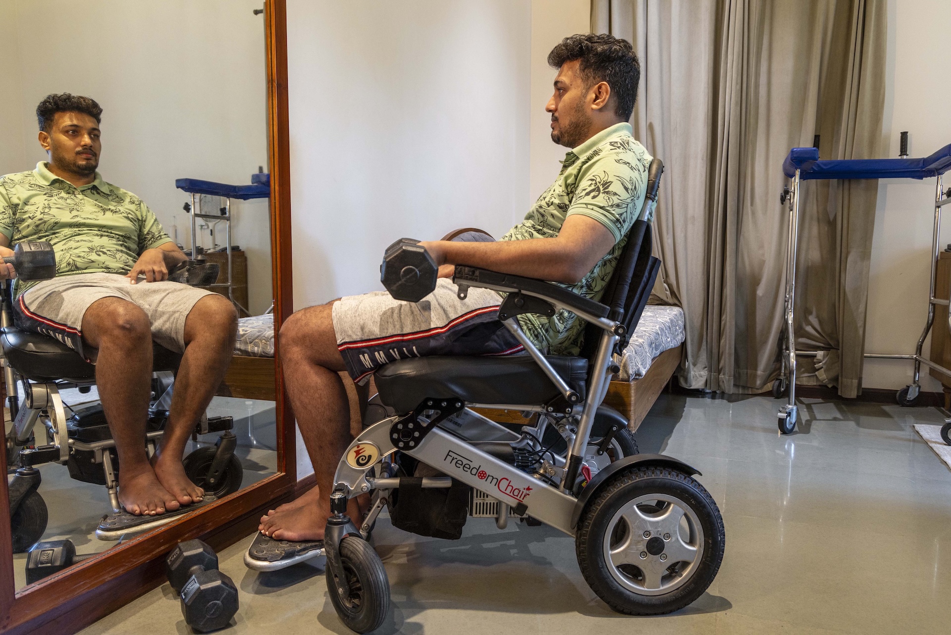 Vais Muhammed (31) sits in a room facing sideways in a silver-coloured motorised wheelchair with black leather seat and arm rests. He is reflected in a large rectangular mirror with a tan wooden frame. His curly hair is short at the sides and he has a thin moustache and a trimmed beard that lines his cheeks and chin. His bare feet rest on the wheelchair’s footrest and he holds a black dumbbell in his left hand with the arm on the arm rest. He wears pale grey shorts and a pista green half-sleeved knitted shirt printed with a black leafy design.