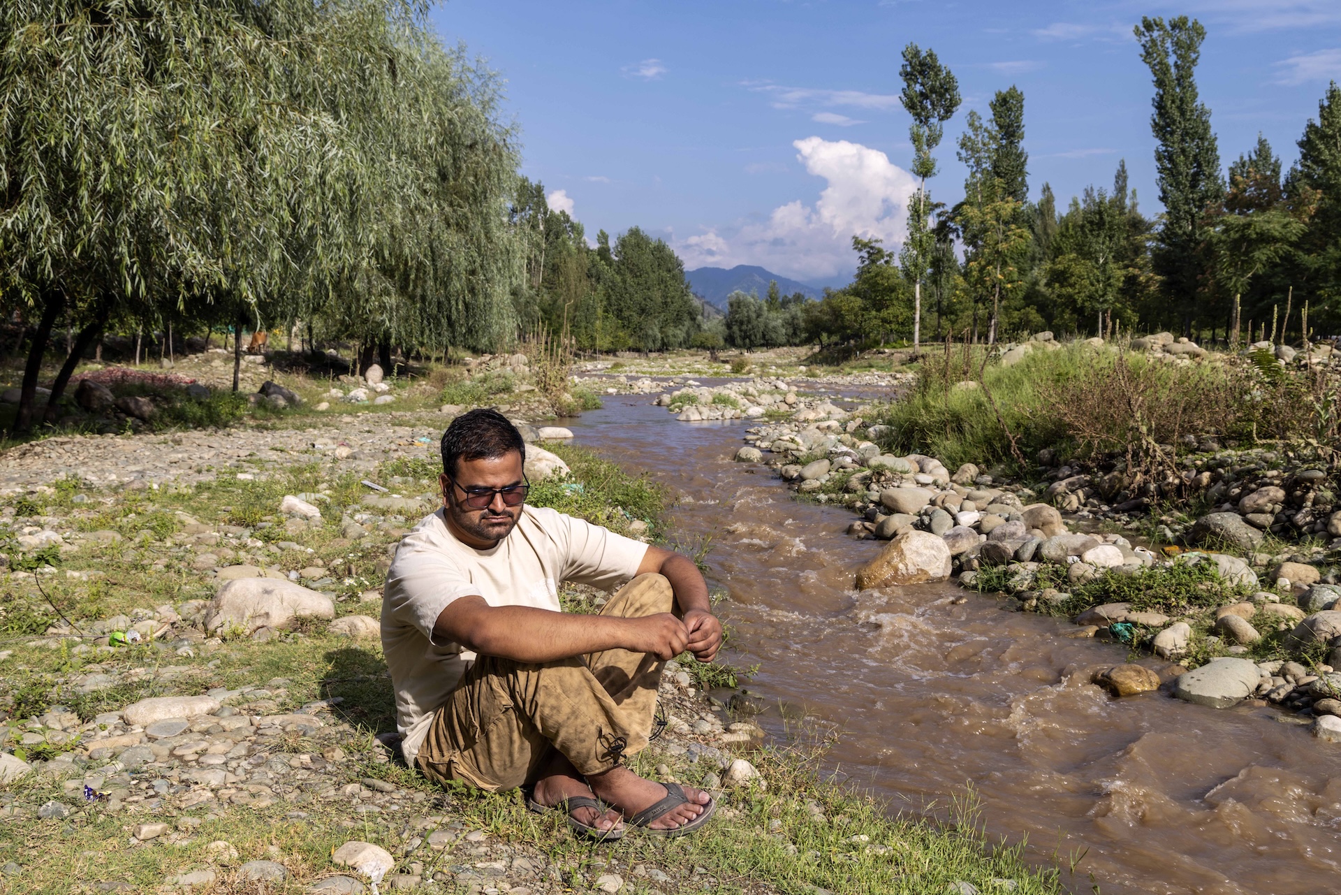 Yasir Ahmad Khan (29) sits on the verge of a flowing mud-brown rivulet that stretches into the distance. There are green trees, mainly pines and willows, in the background. The ground on which Yasir sits has stubbly green grass growing around small white smooth rocks and pebbles. The other side of the narrow rivulet has slightly larger beige and pale brown rocks smoothened by the water. Yasir has short black hair with a side parting, a thin moustache and a stubbly beard lining his cheeks and chin. He wears tinted glasses, a loose cream T-shirt, and loose mocha brown pants. He sits with arms resting on his folded knees.