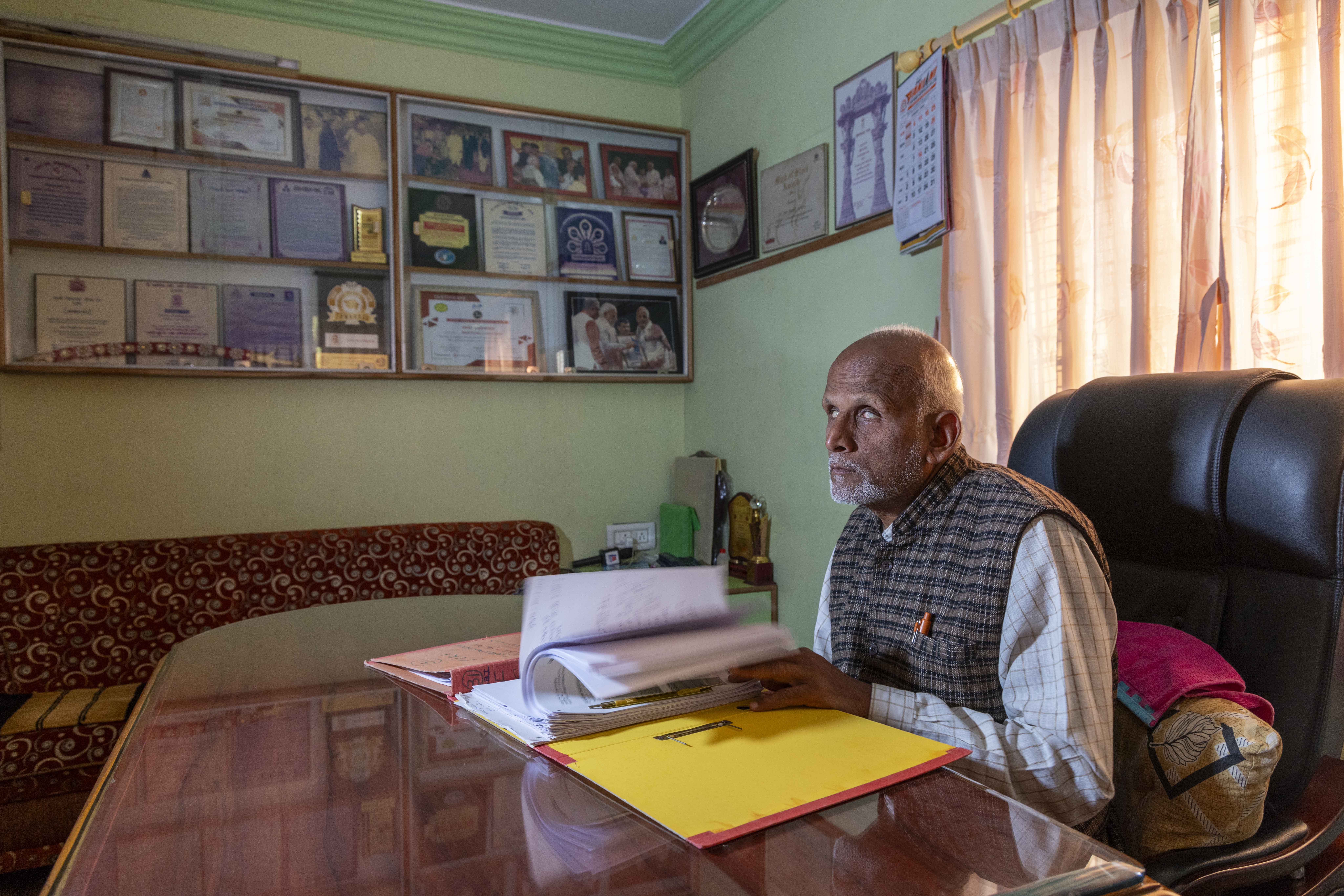 Side shot of Yusufi Kapadiya (62) sitting in a black stuffed leather chair behind a table covered with a clear glass sheet. Sunlight filters through thin salmon pink window curtains behind the chair. Yusufi’s head is bald on top and fringed with white hair. He has a close-shaven, sparse white beard and moustache. He wears a half-sleeved vest of black plaid checks on a pale brown background, over a full-sleeved shirt with graph checks of black on white. His hands are rifling the white sheets of a box-file in front of him. To its right is a closed box file with a pink cover. Fixed to the cream wall behind the table there are two large rectangular frames of tan-coloured wood. Within these frames, certificates framed in the same wood have been arranged in three layered rows.