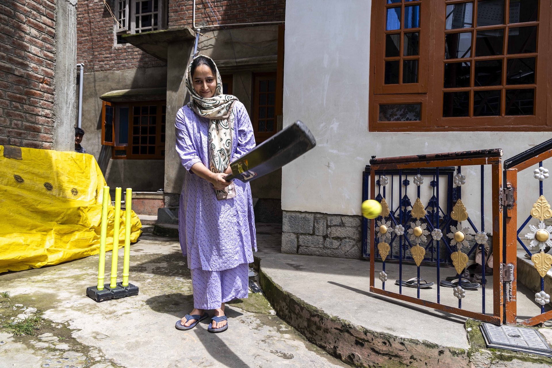 Zakiya Manzoor (24) smiles as she stands on a rough concrete surface, holding a dark brown cricket bat in both hands and raising it to waist-level preparing to strike. A yellow ball is mid-air. Three yellow plastic stumps on a black plastic base form the wicket. Zakiya wears a lilac salwar-kameez printed with tiny black dots. A beige dupatta edged in brown and printed with black swirly motifs is wrapped around her head and neck with one end falling down her chest. She has ash-grey slippers on her feet. There is the façade of a building behind her, made of red exposed bricks.